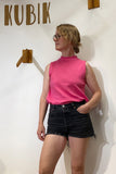 Wearing the Kubik top cineast pink by BUTIK KUBIK—a 60’s inspired, A-line sleeveless jersey top in pink—paired with black denim shorts, a person stands before a white wall featuring "KUBIK" above and wooden hooks beside them, gazing left with one hand tucked in their pocket.