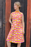 A woman stands before dark wooden doors, wearing the BUTIK KUBIK Kubik dress Twist with Cineast combo Water Lily—a 50s-inspired jersey dress featuring a high collar and a colorful floral pattern. She gazes to the side with one hand in her pocket.