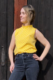 A woman stands before a rustic wooden door, wearing the BUTIK KUBIK Kubik top cineast polkadot yellow with high-waisted dark jeans. She gazes to the side, smiling slightly with her hand on her hip.