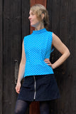 A person stands in front of a dark wooden door, wearing the Kubik top cineast polkadot turquoise from BUTIK KUBIK with a short black skirt and a front zipper. They pose with one hand on their hip, looking to the side.