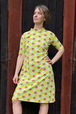 A woman poses confidently before a wooden wall in the BUTIK KUBIK Kubik dress cinema midi krokus—a 60's-inspired light green midi dress with vibrant pink and yellow floral patterns.