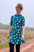 A woman with glasses stands outdoors on a dirt path, wearing the Kubik dress cinema anemone green by BUTIK KUBIK. Grassy fields and trees are in the background on a cloudy day.