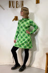 A person poses in front of a white wall with "KUBIK" above, wearing the BUTIK KUBIK Kubik dress cinema klöver lime— a 60's-inspired A-line long sleeve dress with shamrock pattern—paired with black leggings and shoes.