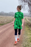 A woman wears the BUTIK KUBIK Kubik dress cinema the leaf, a green dress with a bold leaf pattern, paired with navy leggings, striped socks, and orange shoes. Her hair is in a bun as she walks along a misty rural dirt road.