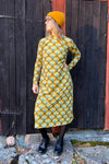 A person stands in front of a dark wooden wall, showcasing the BUTIK KUBIK Kubik dress cinema bridges maxi, featuring a 60s inspired yellow and green geometric design. Accessorized with a yellow beanie, glasses, black tights, and black shoes, they glance to the side.