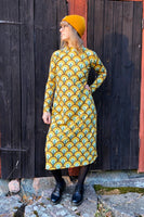 A person stands in front of a dark wooden wall, showcasing the BUTIK KUBIK Kubik dress cinema bridges maxi, featuring a 60s inspired yellow and green geometric design. Accessorized with a yellow beanie, glasses, black tights, and black shoes, they glance to the side.