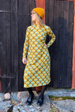 A person stands in front of a dark wooden wall, showcasing the BUTIK KUBIK Kubik dress cinema bridges maxi, featuring a 60s inspired yellow and green geometric design. Accessorized with a yellow beanie, glasses, black tights, and black shoes, they glance to the side.