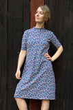 Wearing the Kubik dress cinema midi petite fleur by BUTIK KUBIK, a person poses in front of a dark wooden wall with one hand on their hip, looking to the side and smiling slightly.