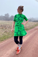 A person in glasses stands on a dirt road wearing the BUTIK KUBIK Kubik dress cinema the leaf, black leggings, striped white socks, and orange shoes, with a grassy field and trees in the misty background.