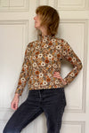 A person with short, light brown hair models the Kubik sweater gerbera by BUTIK KUBIK, made from GOTS certified cotton, paired with dark jeans, standing with one hand on their hip before a paneled white wall.