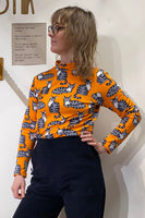 A person with shoulder-length hair and glasses wears the BUTIK KUBIK Kubik sweater caturday, a 60s-inspired orange knit top with a cat pattern, paired with dark pants. They stand against a white wall, with a small wooden board featuring text in the background.