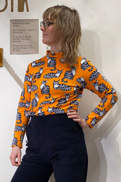 A person with shoulder-length hair and glasses wears the BUTIK KUBIK Kubik sweater caturday, a 60s-inspired orange knit top with a cat pattern, paired with dark pants. They stand against a white wall, with a small wooden board featuring text in the background.