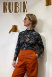 A person wearing glasses poses by a wall with wooden hooks and the word “KUBIK” displayed above, styled in orange pants and the BUTIK KUBIK “Kubik sweater cinema birds,” layered under a floral long-sleeve shirt for a 60’s-inspired look.