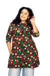 A woman with curly hair smiles wearing the BUTIK KUBIK Cissi och Selma dress/tunic Hassel Rooibos with a colorful floral print and three-quarter sleeves, paired with blue jeans, standing against a white background.