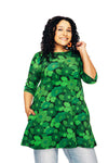 A woman with curly hair smiles, wearing the Cissi och Selma dress/tunic Anita 1970 Mörkgrön from BUTIK KUBIK, featuring a green floral print and three-quarter sleeves, paired with blue jeans and posing against a white background.