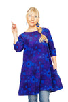 A smiling woman with blond hair in a side braid wears the vibrant BUTIK KUBIK Cissi och Selma dress/tunic Anita 1970 Violett, paired with jeans, standing and pointing upward with her right hand against a white background.