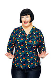 A smiling woman with a short black bob wears the Cissi och Selma blus Susanne Droppe Mörkblå by BUTIK KUBIK, featuring a colorful geometric pattern. She poses cheerfully with raised hands and spread fingers against a white background.