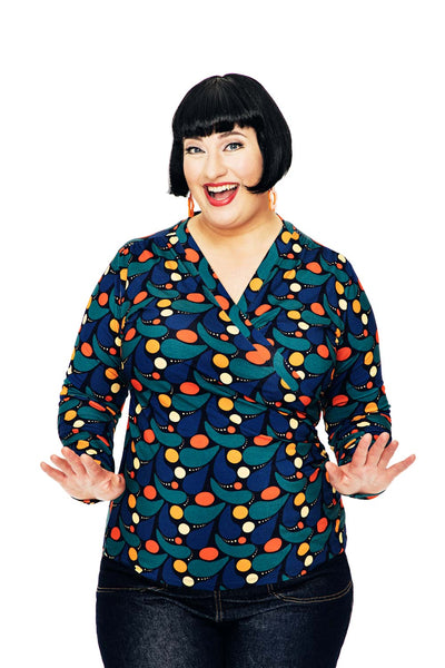 A smiling woman with a short black bob wears the Cissi och Selma blus Susanne Droppe Mörkblå by BUTIK KUBIK, featuring a colorful geometric pattern. She poses cheerfully with raised hands and spread fingers against a white background.