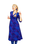 A smiling woman with long blond braided hair wears the BUTIK KUBIK Cissi och Selma dress Vera 1970 Violett, paired with matching earrings, standing with both index fingers raised against a white background.