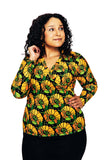 A woman with curly hair stands against a white background, wearing the Cissi och Selma blus Susanne Kronblad Senap from BUTIK KUBIK—a colorful, long-sleeved blouse—and a black skirt. She smiles gently and gazes slightly to her left.
