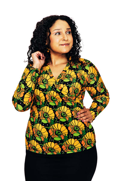A woman with curly hair stands against a white background, wearing the Cissi och Selma blus Susanne Kronblad Senap from BUTIK KUBIK—a colorful, long-sleeved blouse—and a black skirt. She smiles gently and gazes slightly to her left.