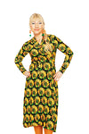 A woman with blonde hair in a side braid wears the BUTIK KUBIK Cissi och Selma Anna-Klara dress Kronblad Senap, orange earrings, and orange tights, standing with hands on hips against a white background.