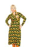 A woman with blonde hair in a side braid wears the BUTIK KUBIK Cissi och Selma Anna-Klara dress Kronblad Senap, orange earrings, and orange tights, standing with hands on hips against a white background.
