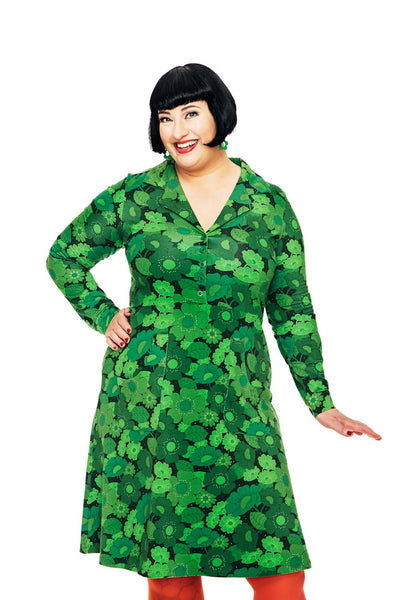 A smiling woman with short black hair wears the BUTIK KUBIK Cissi och Selma dress Viveca 1970 Mörkgrön in eco-friendly cotton jersey and orange leggings, standing against a white background with one hand on her hip.