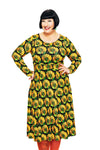 A smiling woman with short black hair stands with hands on hips, wearing the Cissi och Selma dress Helga Kronblad Senap by BUTIK KUBIK and orange tights, against a white background.