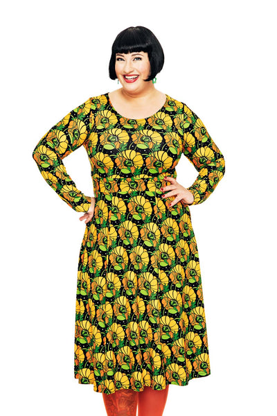 A smiling woman with short black hair stands with hands on hips, wearing the Cissi och Selma dress Helga Kronblad Senap by BUTIK KUBIK and orange tights, against a white background.