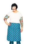 A smiling woman with short black hair and colorful tattoos wears a white top, the BUTIK KUBIK Cissi och Selma Majken skirt Miniblom Blå, teal tights, and bright lipstick against a white background.