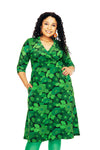 A woman with long curly hair smiles while wearing the Cissi och Selma dress Vera 1970 Mörkgrön by BUTIK KUBIK, a green floral dress with three-quarter sleeves and green tights, standing with one hand in her pocket against a white background.