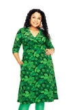 A woman with long curly hair smiles while wearing the Cissi och Selma dress Vera 1970 Mörkgrön by BUTIK KUBIK, a green floral dress with three-quarter sleeves and green tights, standing with one hand in her pocket against a white background.