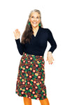 A smiling woman with long gray hair, in a black top, BUTIK KUBIKs Cissi och Selma Majken skirt Rooibos with colorful florals, orange tights, and green earrings poses playfully with one hand raised against a white background.