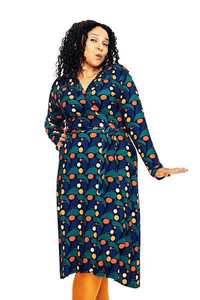 A woman with curly dark hair poses confidently in the BUTIK KUBIK Cissi och Selma Anna-Klara dress Droppe Mörkblå, featuring red, orange, and yellow shapes, paired with yellow tights against a plain white background.