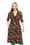 A smiling woman with long gray hair models the Cissi och Selma dress Vera Hassel Rooibos by BUTIK KUBIK, a knee-length, V-neck dress with a bold, colorful floral pattern. She stands against a white background with her hands in her pockets.