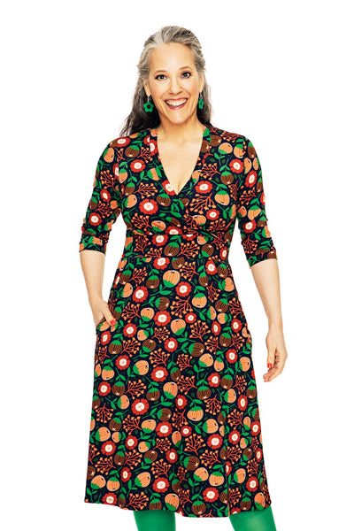 A smiling woman with long gray hair models the Cissi och Selma dress Vera Hassel Rooibos by BUTIK KUBIK, a knee-length, V-neck dress with a bold, colorful floral pattern. She stands against a white background with her hands in her pockets.