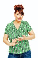 A beehive-styled redhead smiles and gestures, wearing the Cissi och Selma Margit blus Brygga from BUTIK KUBIK in a 50s graphic green design with blue jeans against a white background.