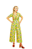 A woman wearing a bright yellow floral Cissi och Selma Britta jumpsuit by BUTIK KUBIK stands confidently with hands on hips. Her long braided hair complements the eco-friendly, ekologisk bomullstrikå fabric. She wears brown shoes and smiles slightly against the white background, showcasing her colorful outfit.