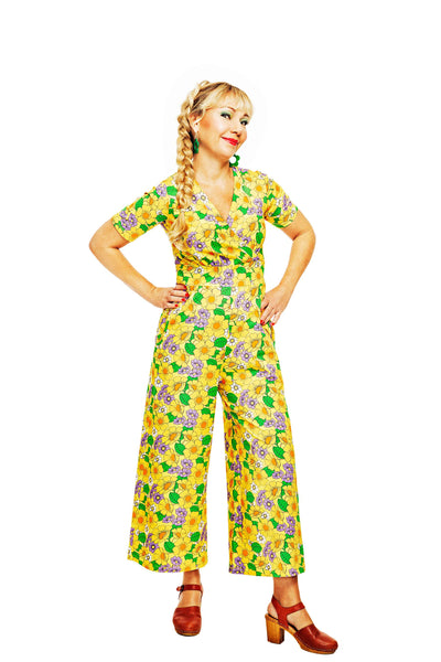 A woman wearing a bright yellow floral Cissi och Selma Britta jumpsuit by BUTIK KUBIK stands confidently with hands on hips. Her long braided hair complements the eco-friendly, ekologisk bomullstrikå fabric. She wears brown shoes and smiles slightly against the white background, showcasing her colorful outfit.