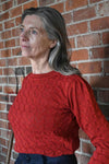 A woman with long gray hair wears a BUTIK KUBIK Palava Clara Rust Pointelle top and stands pensively in front of a brick wall, looking to the side.