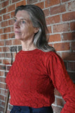 A woman with long gray hair wears a BUTIK KUBIK Palava Clara Rust Pointelle top and stands pensively in front of a brick wall, looking to the side.