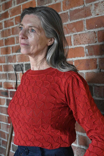 A woman with long gray hair wears a BUTIK KUBIK Palava Clara Rust Pointelle top and stands pensively in front of a brick wall, looking to the side.