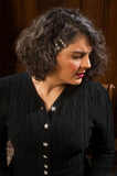 A woman with curly brown hair and red lipstick looks down, wearing the Emmy - The Ice Skater black cardigan by BUTIK KUBIK, featuring vintage style star-shaped buttons and matching star hair clips, set against a wooden-paneled background.