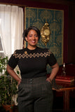 A woman stands indoors, smiling as she wears the Emmy - The Darling Diamond Top in black by BUTIK KUBIK, which features a gold pattern. She pairs it with plaid pants reminiscent of 1930s and 1940s jumpers. Behind her are a gold clock, books, a candle, and green wallpaper with a cream pattern. A leafy plant sits by a window.