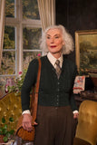 An older woman with white hair stands indoors, smiling as she wears the BUTIK KUBIK Emmy - The Ice Skater cardigan in dark bottle green, featuring a subtle Celtic knit pattern. She has it layered over a white shirt and plaid tie, paired with brown pants, and holds a tan shoulder bag. Vintage decor and greenery serve as the background.