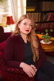 A woman with long red hair sits in a red armchair, wearing the BUTIK KUBIK Emmy - The Ice Skater cardigan in black cherry, layered over her dark sweater and shirt. She is in a cozy room with bookshelves, a table, a potted plant, and sunlight streaming through the window.