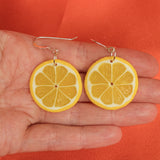 A hand holds a pair of Becca design Citron earrings by BUTIK KUBIK, shaped like lemon slices with yellow rinds and detailed sections, against an orange background.