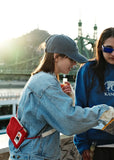 Two people outdoors, one wearing a denim jacket and cap, sporting a red BUTIK KUBIK YKRA travel Red waist bag and compact organizer. As they look at something together with the sunset behind them—framed by a bridge and building—the scene highlights the practicality of the BUTIK KUBIK YKRA travel Red as an essential travel accessory.