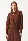 A woman wearing the Jumperfabriken Blouse Emmi Brown by BUTIK KUBIK, featuring a tied bow at the neck and crafted from mjuk kvalitet, paired with a matching pleated skirt. She stands against a plain white background, embodying a modern vintage style.
